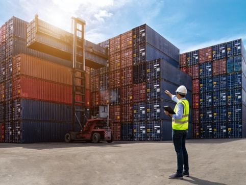 Man with high vis jacket and hard hat with tablet pointing to Shipping containers stacked at a dock